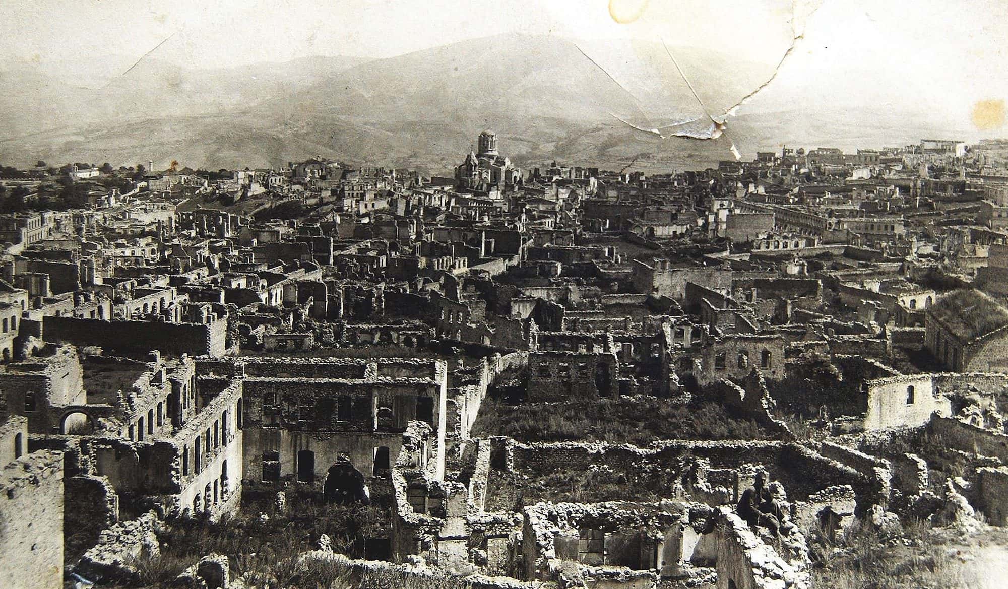Wide black and white view of a destroyed hillside quarter, roofless stone shells, a large domed cathedral standing behind