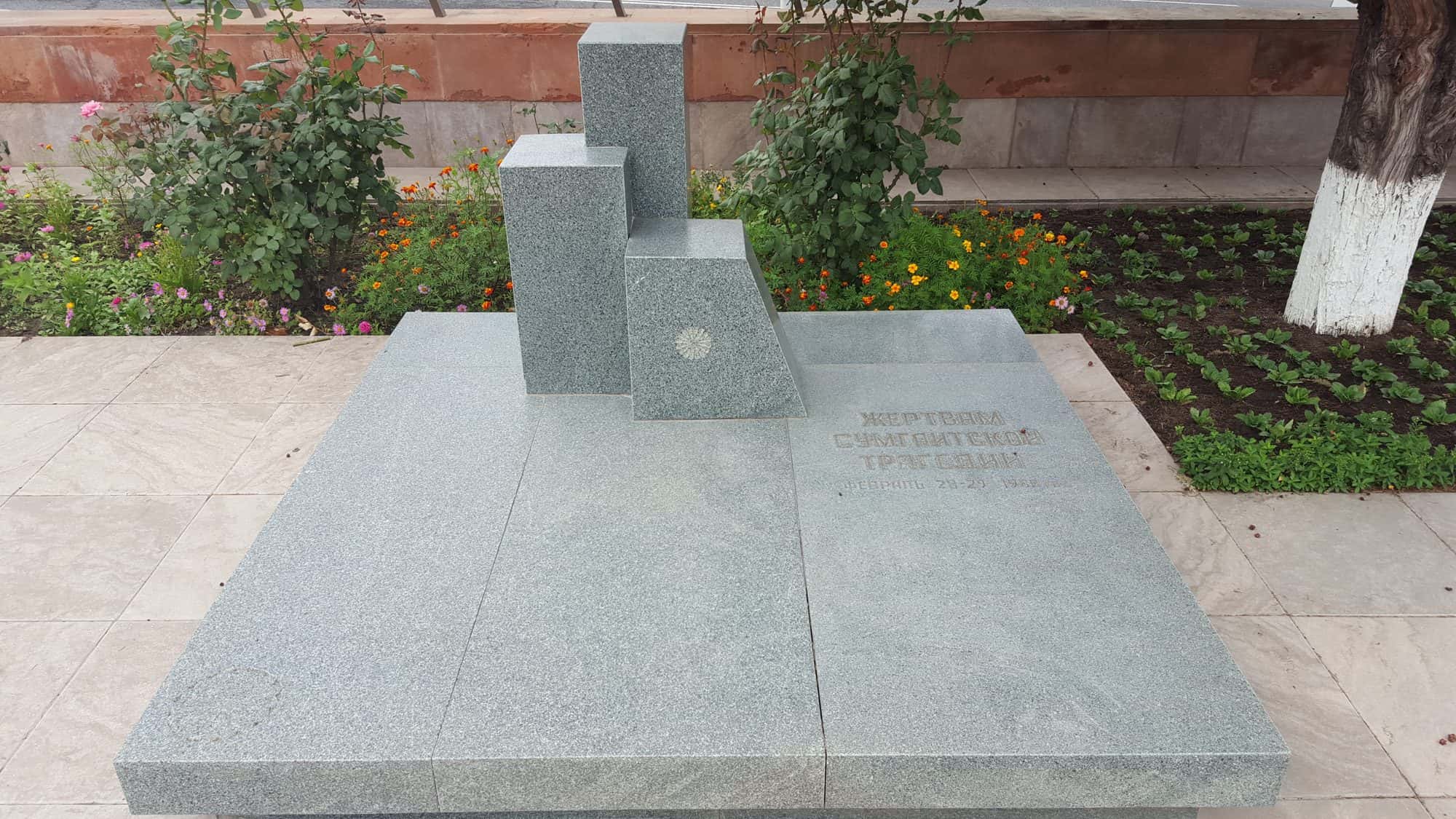 Grey granite memorial of stepped blocks on a paved platform, flowerbeds and trees behind