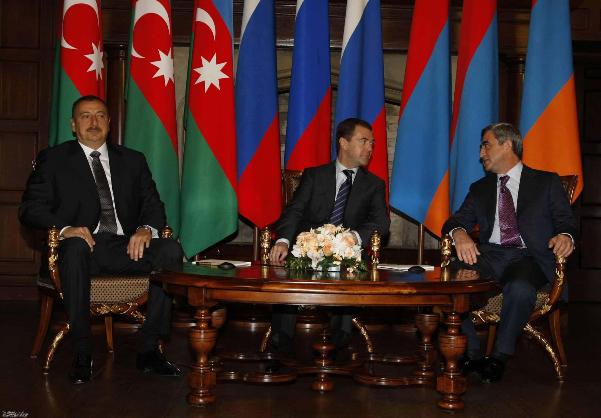 Three heads of state seated at a long table signing documents before national flags