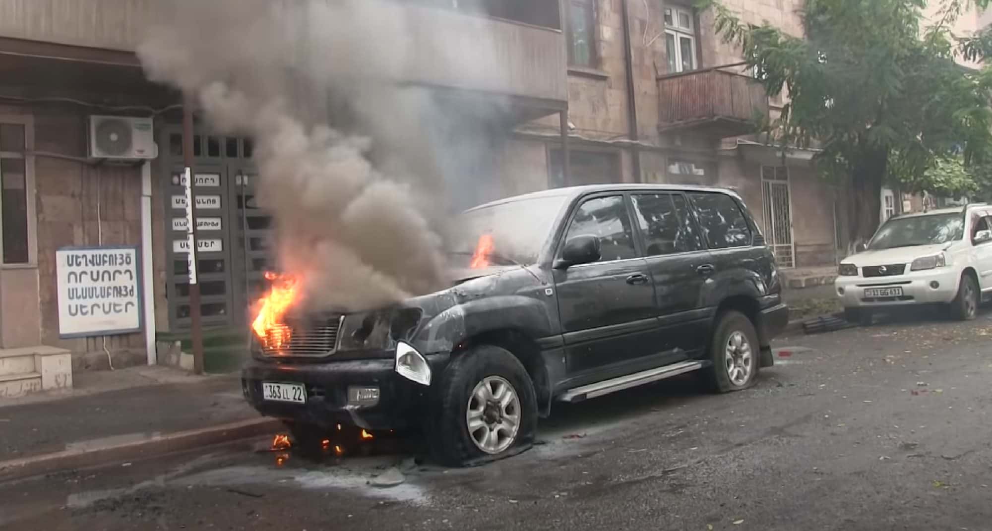 Burning four wheel drive vehicle on a city street with smoke rising past damaged apartment buildings
