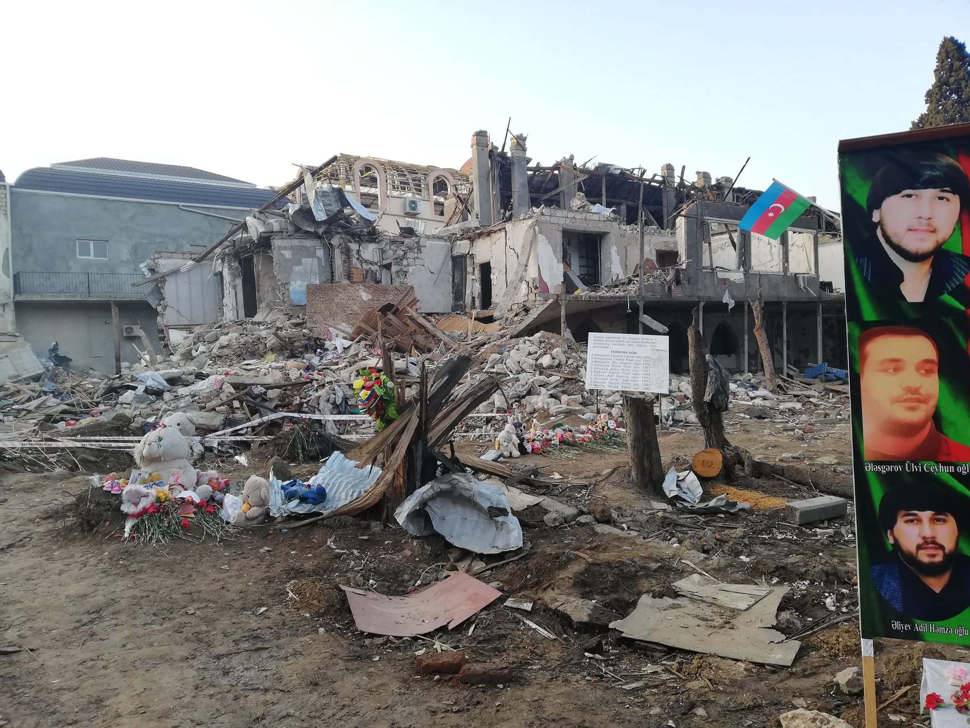Levelled houses and rubble at a bomb site, with flowers and a banner of portraits placed as a memorial