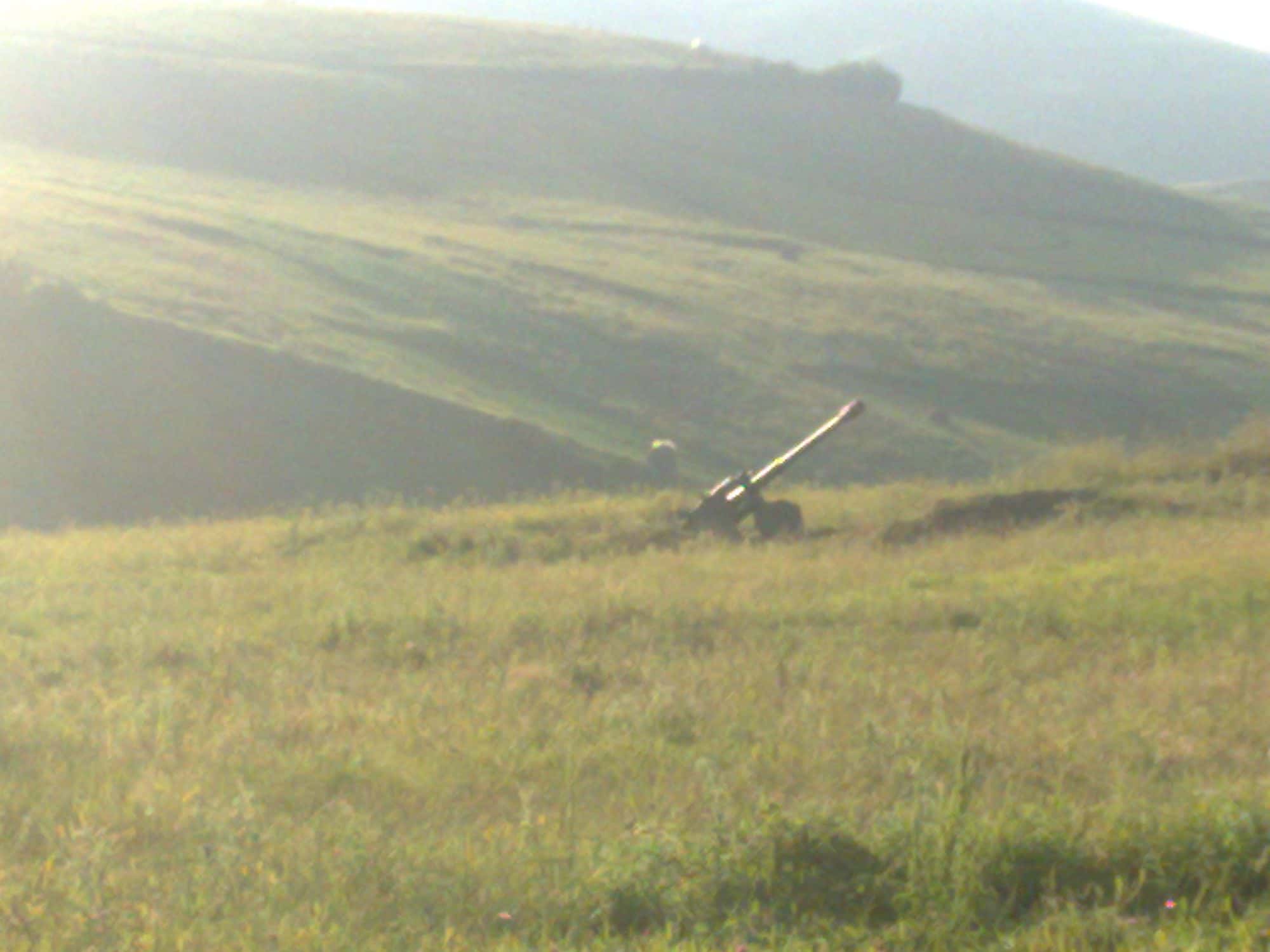 Camouflaged artillery piece positioned on a green hillside near a village on the border