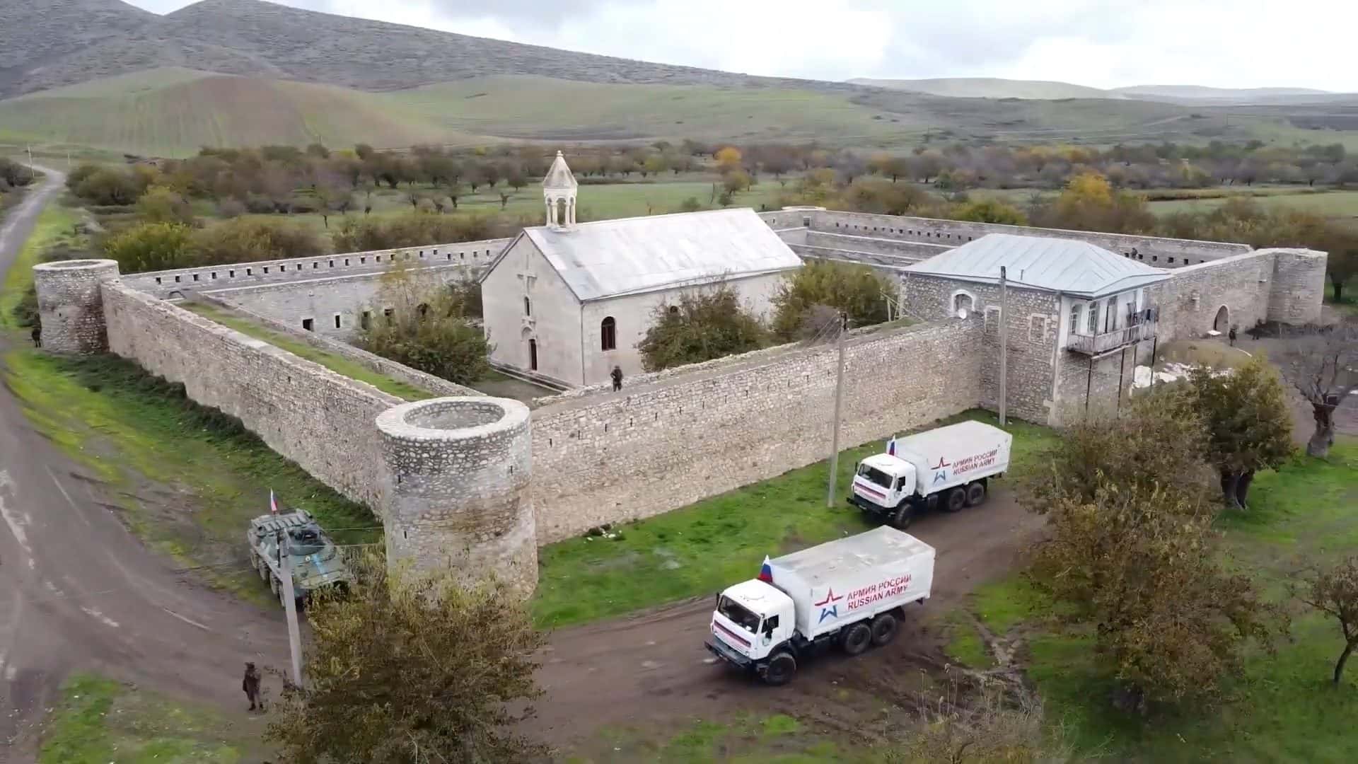 Aerial view of a walled stone monastery with two military trucks parked outside its gate