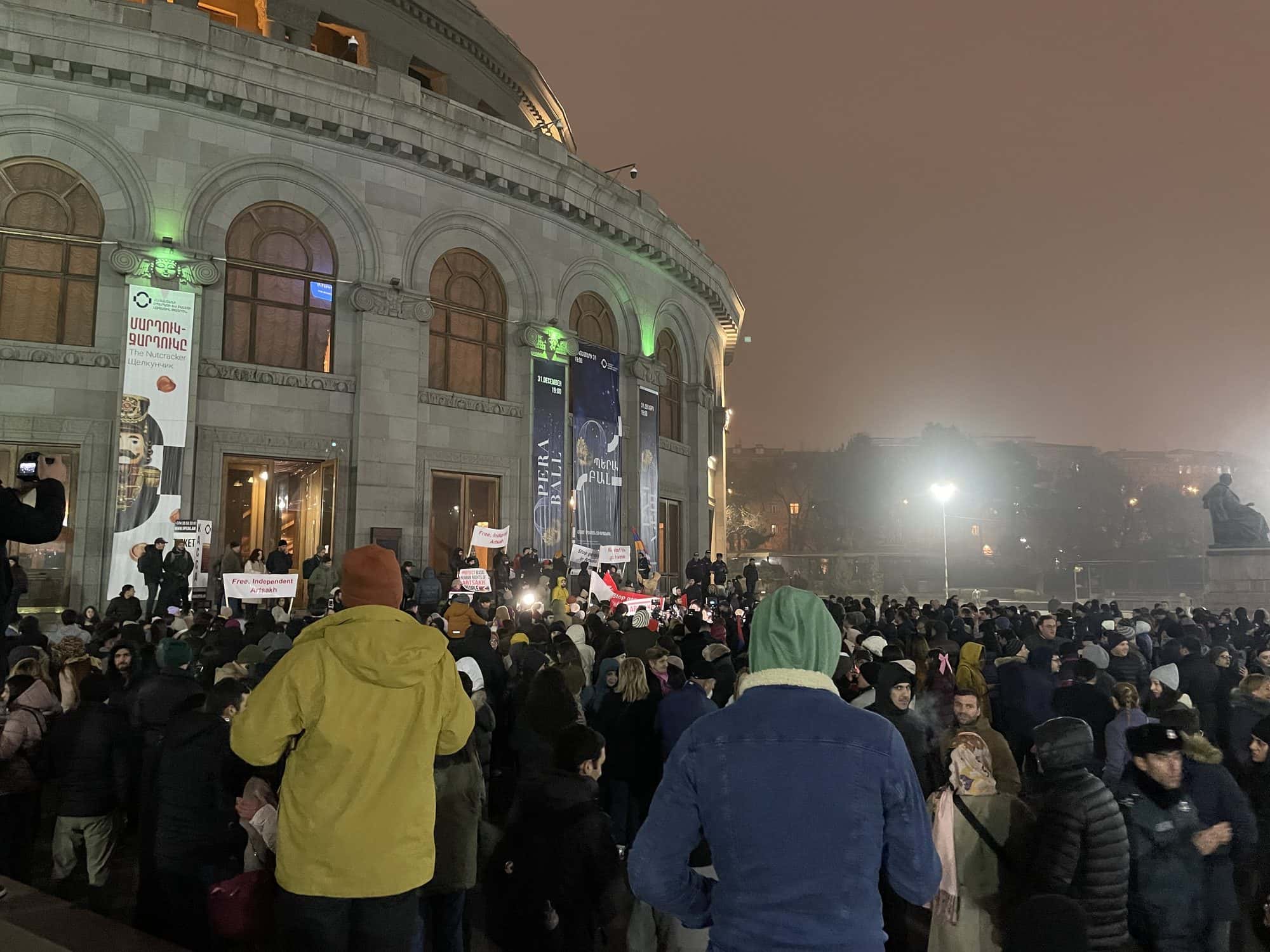 Large night time crowd filling a city square in front of a lit opera house
