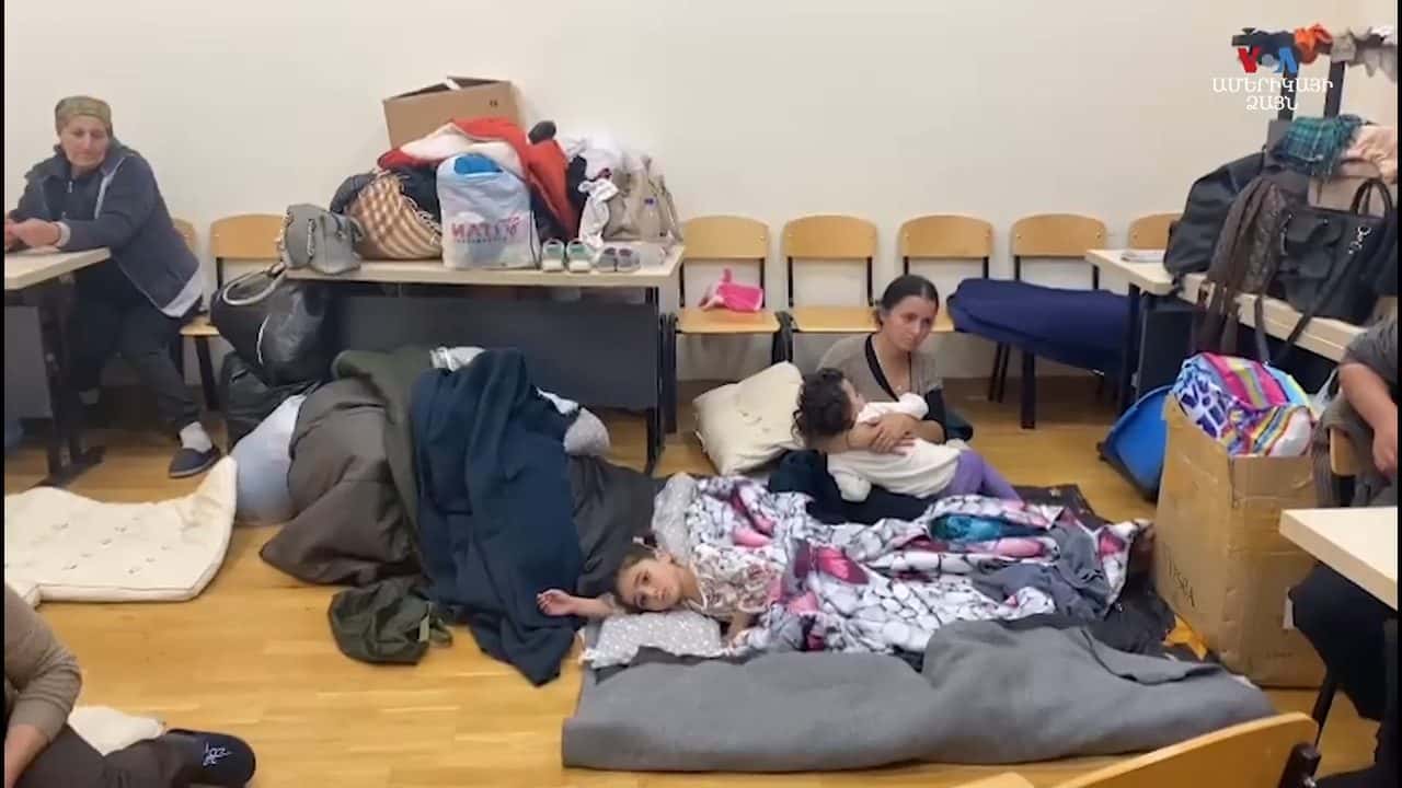 Displaced families resting on bedding on a hall floor with suitcases and bundles stacked around