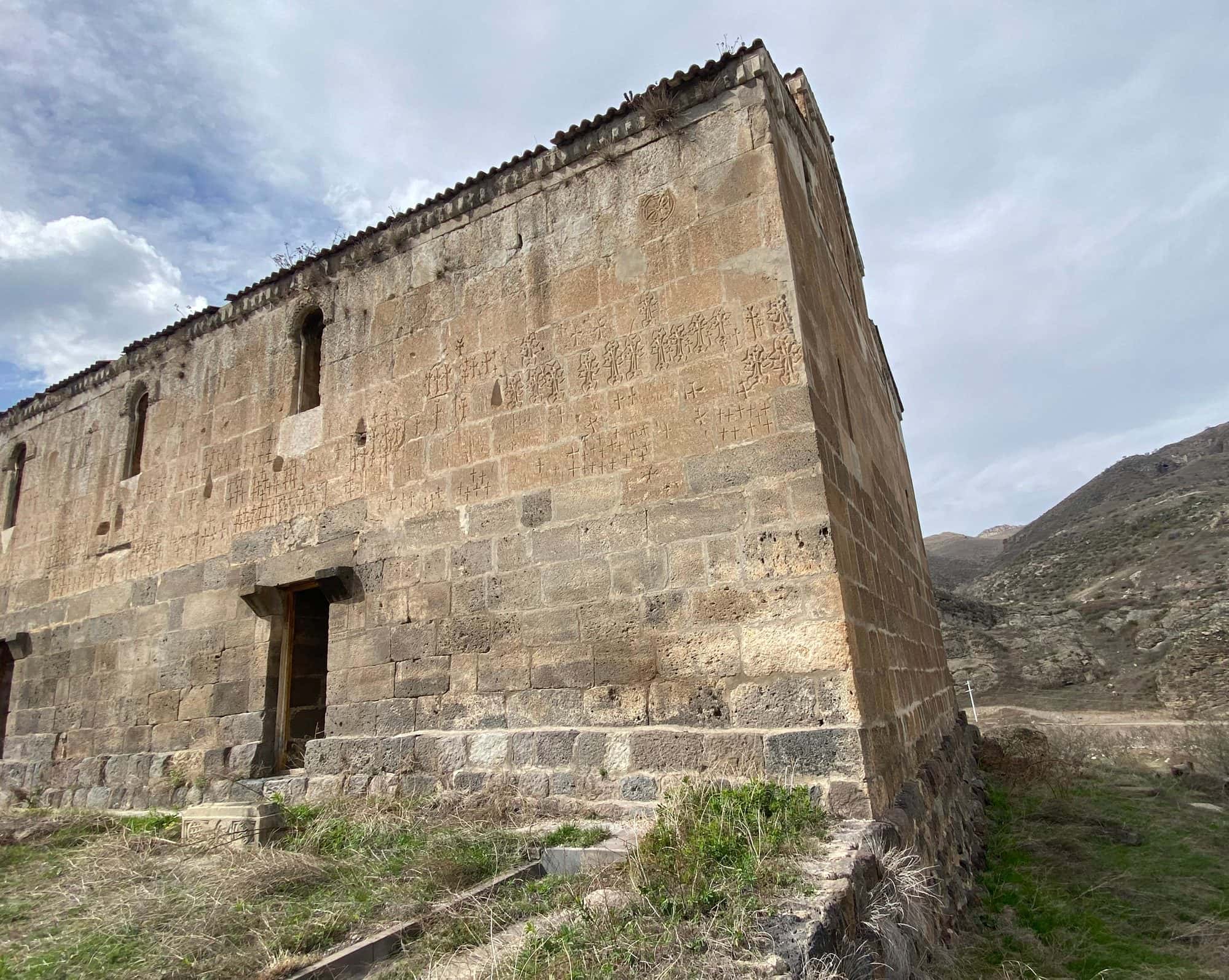 Ancient stone basilica with a tiled gable roof and small windows, standing in a grassy walled yard