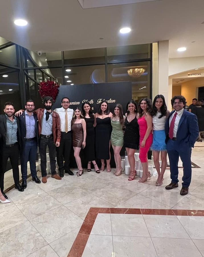Twelve ARF Shant members in formal dress, arms around each other, in front of an Armenian Youth Federation banner at an evening event.