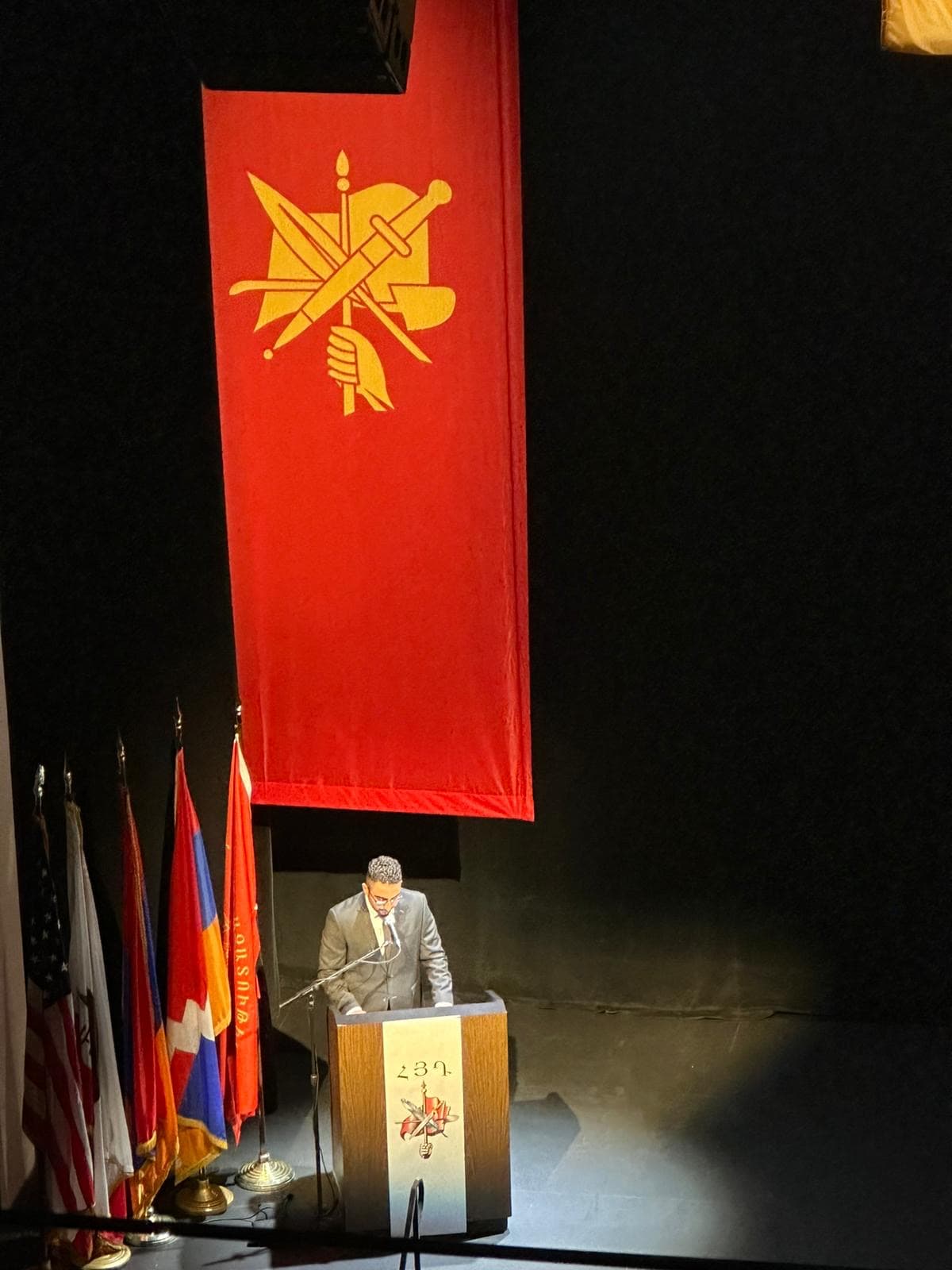 A speaker at a lectern on a darkened stage, beneath the ARF's banner of the crossed sword, pen and spade, with the American, Armenian and Artsakh flags standing beside him.