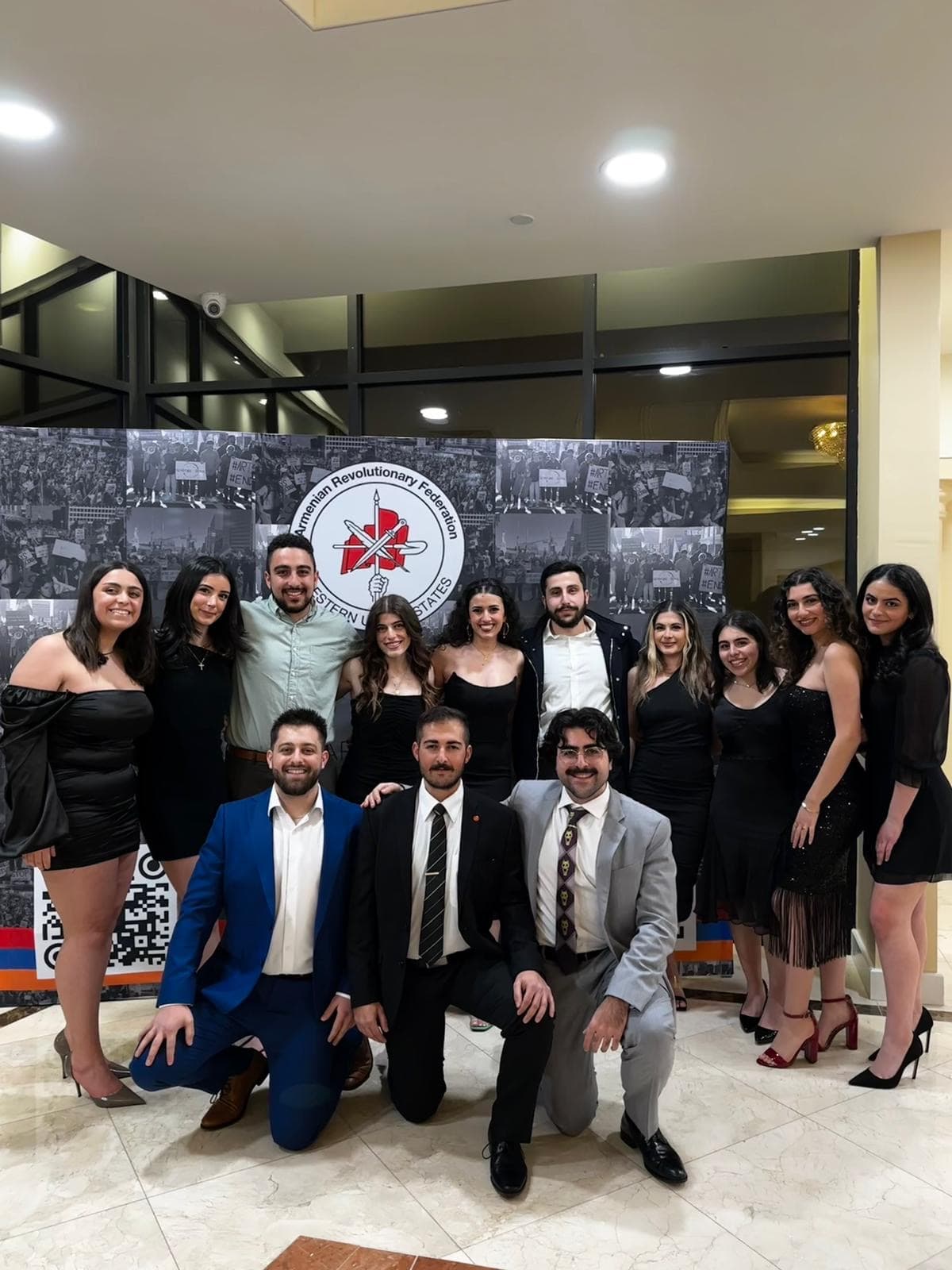 Fourteen ARF Shant members posed together, three kneeling in front, against a backdrop of protest photographs and the seal of the Armenian Revolutionary Federation Western United States.