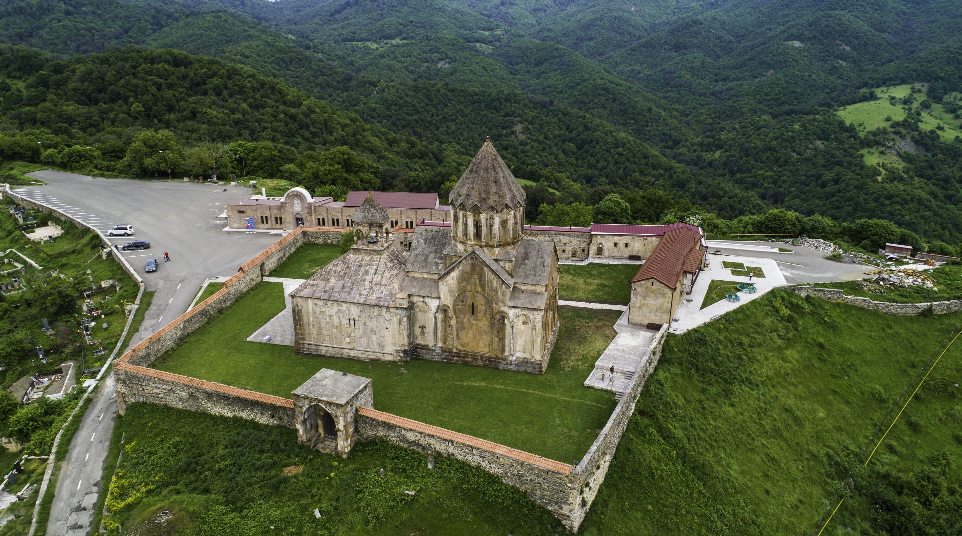 Gandzasar Monastery, seen from the air: ochre stone church inside a walled compound on a green ridge, forested hills behind it